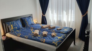 A neatly made bed with blue bedding and rolled towels, a bedside lamp, and a view of the outside through sheer curtains.