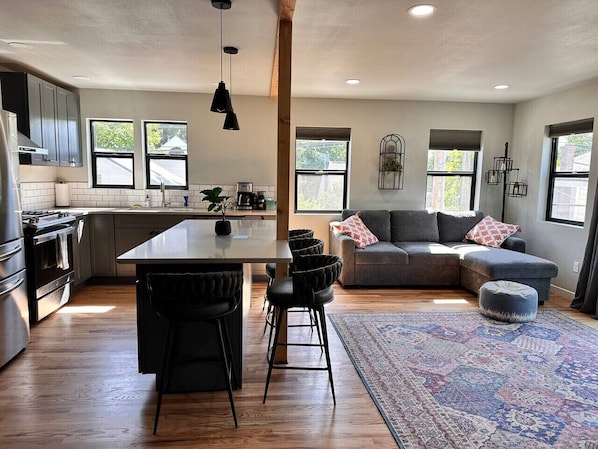 A modern kitchen with a central island, bar stools, and a grey sofa in a living area with a patterned rug.