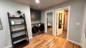 A laundry room with two front-loading washers and dryers, a wooden floor, and a dark metal shelving unit with a plant on top.