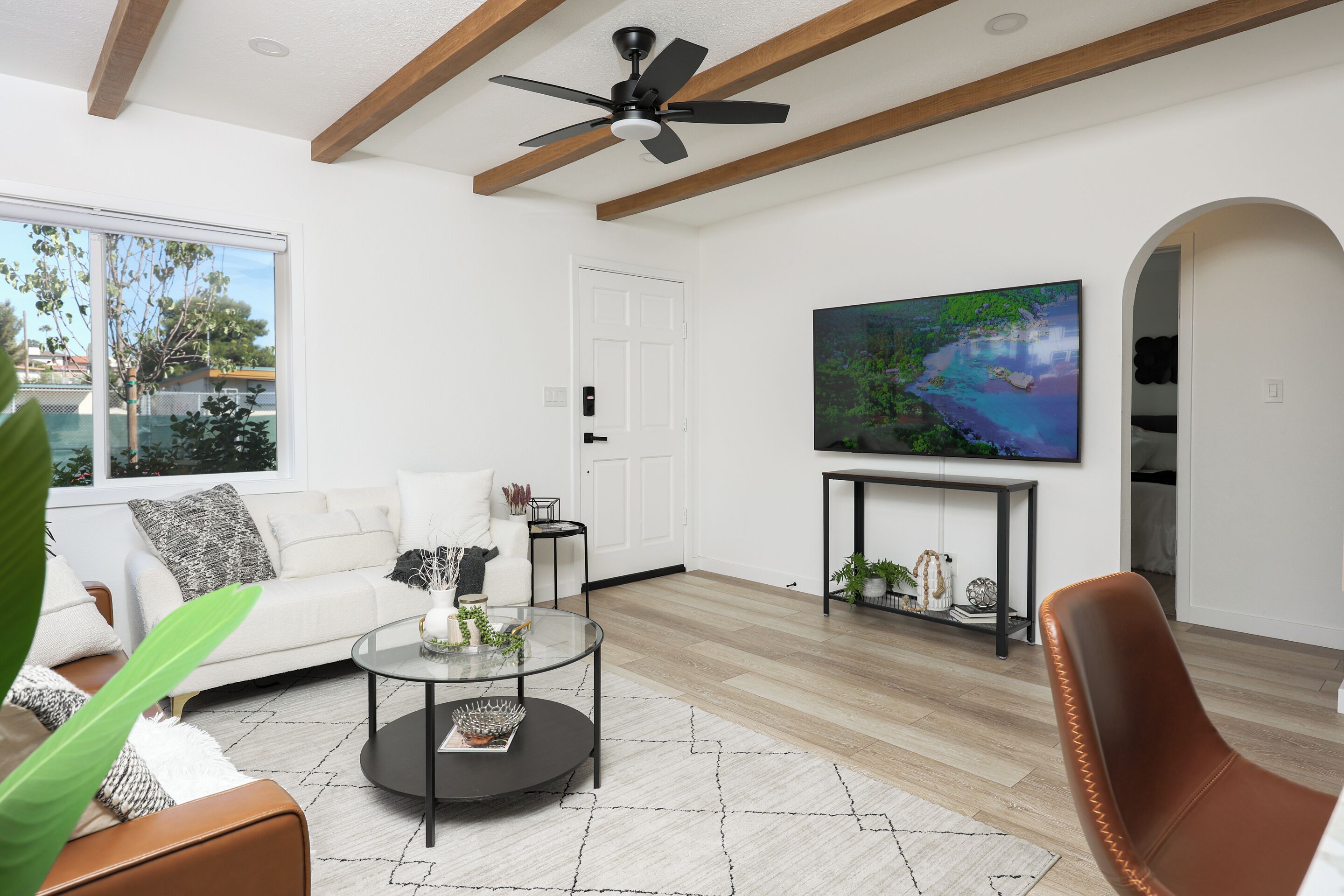 A modern living room with a white sofa, a glass coffee table, a brown leather chair, a ceiling fan, and a large TV on a black console.