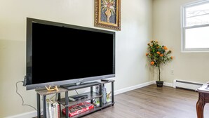 A living room with a large flat-screen TV on a wooden stand, a small bookshelf, and a potted plant with orange flowers. - 2 Mi to The Marketplace: Spring Valley Apartment (Spring Valley)