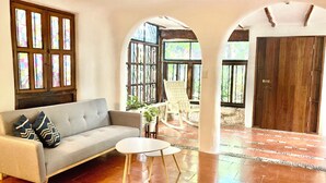 A living room with a sofa, two small tables, and a rocking chair. There are large windows with stained glass, a door, and a tiled floor. - Cozy apartment in tropical villa surrounded by trees. Oasis in the city! (Cancún)