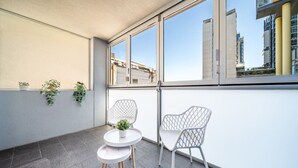 A balcony with two white chairs and a small table, potted plants, and a view of buildings through large windows.