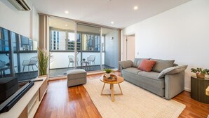 A modern living room with a grey sofa, a flat-screen TV, a wooden coffee table, and a large sliding glass door leading to an outdoor balcony.