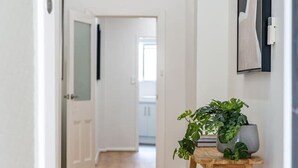 Un couloir au sol en bois, une plante en pot posée sur un tabouret et une porte donnant sur une autre pièce.