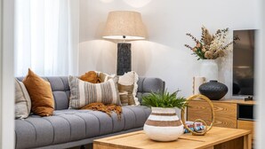 Un salon moderne avec un canapé gris, une table basse en bois, une lampe, un vase contenant des fleurs séchées et un téléviseur posé sur un meuble en bois.