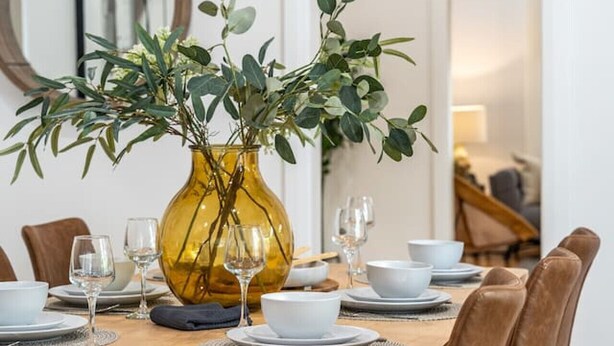 Une table à manger dressée avec des assiettes, des verres et un vase jaune orné de feuillage vert.