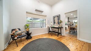Un salon moderne avec un plancher en bois, un tapis gris, un fauteuil noir agrémenté de coussins colorés, une petite table sur laquelle est posée une plante, et une œuvre d’art encadrée accrochée au mur.