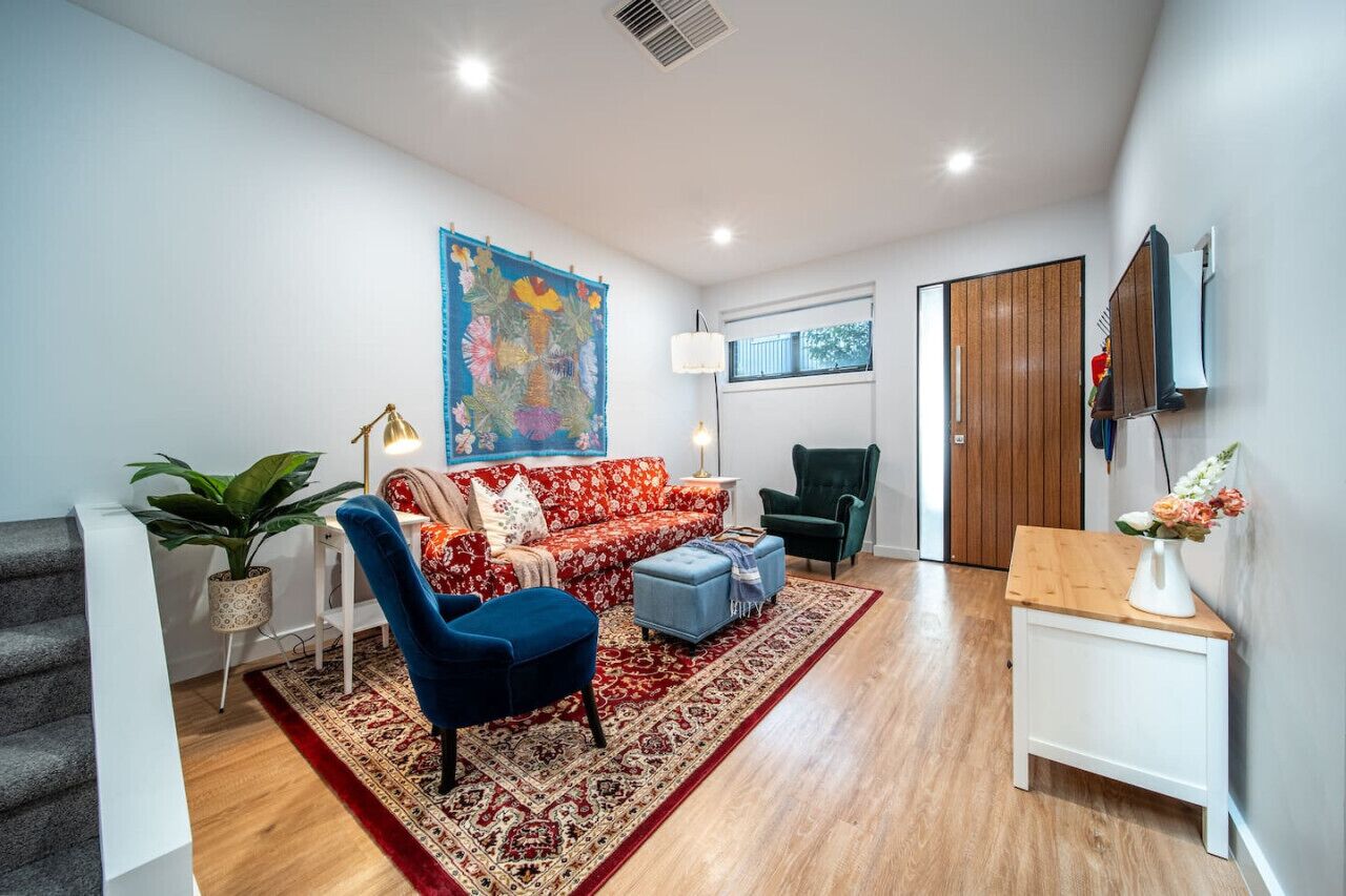 Un salon moderne avec un canapé à motifs floraux rouges, un fauteuil bleu et une table basse en bois. Un grand tableau abstrait orne le mur, et une plante en pot se trouve sur la gauche.