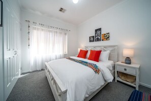 Une chambre avec un lit blanc, des oreillers rouges, une table de chevet avec une lampe et des œuvres d’art accrochées au mur.