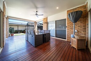 Une terrasse extérieure couverte avec une plateforme en bois, une table à manger avec des chaises, un ventilateur de plafond et une grande porte-fenêtre coulissante.
