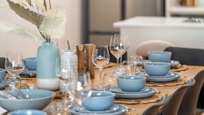 Ein Esstisch ist gedeckt mit blauen Schüsseln, Tellern und Weingläsern, ergänzt durch eine Vase mit weißen Federn und einer strukturierten Vase.