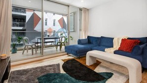 Ein modernes Wohnzimmer mit einem blauen Sofa, einer weißen Bank und einer Glastür, die auf einen Balkon mit Esstisch und Stühlen führt.