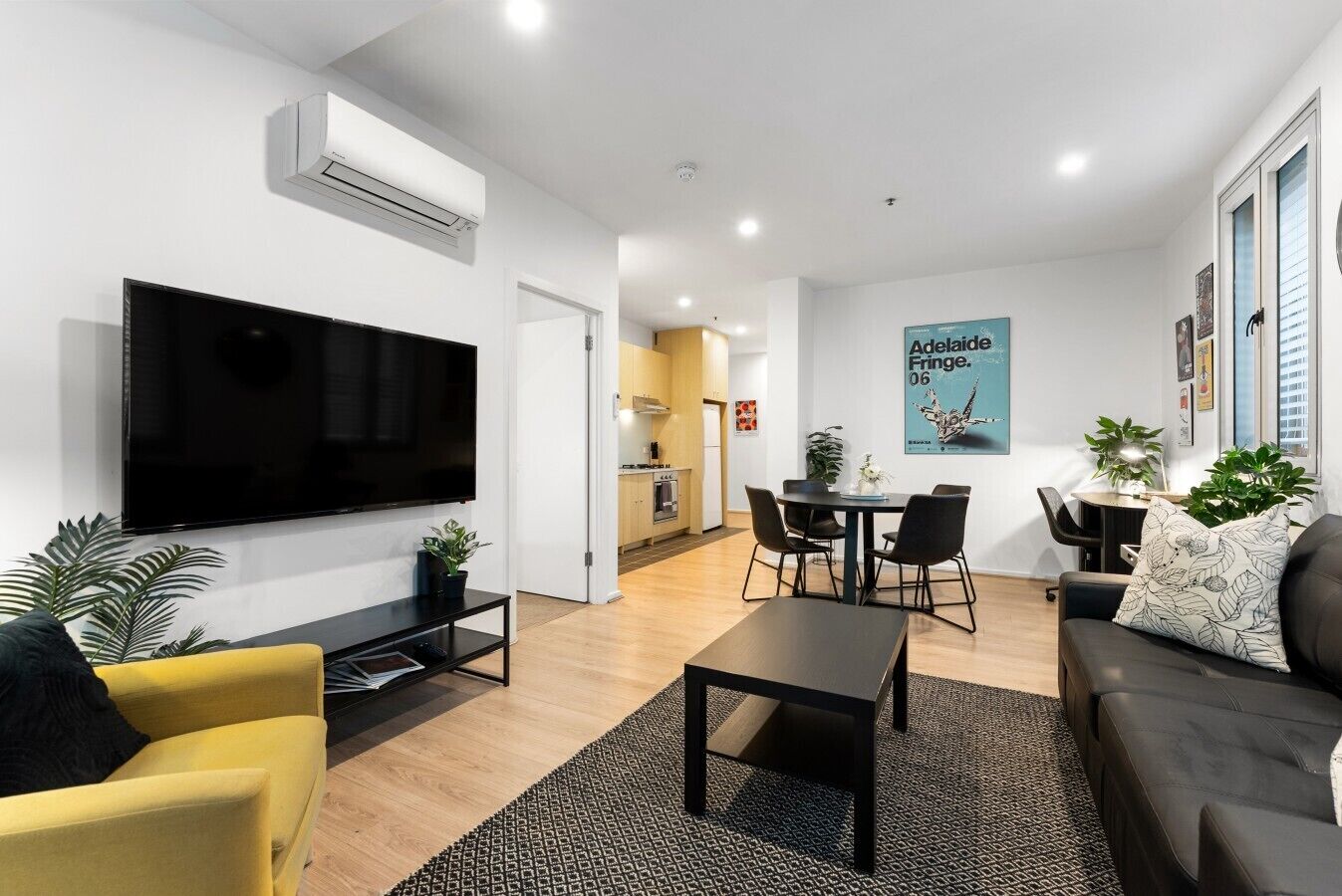 A modern living room with a flat-screen TV, a yellow armchair, a black sofa, and a dining area with a table and chairs.