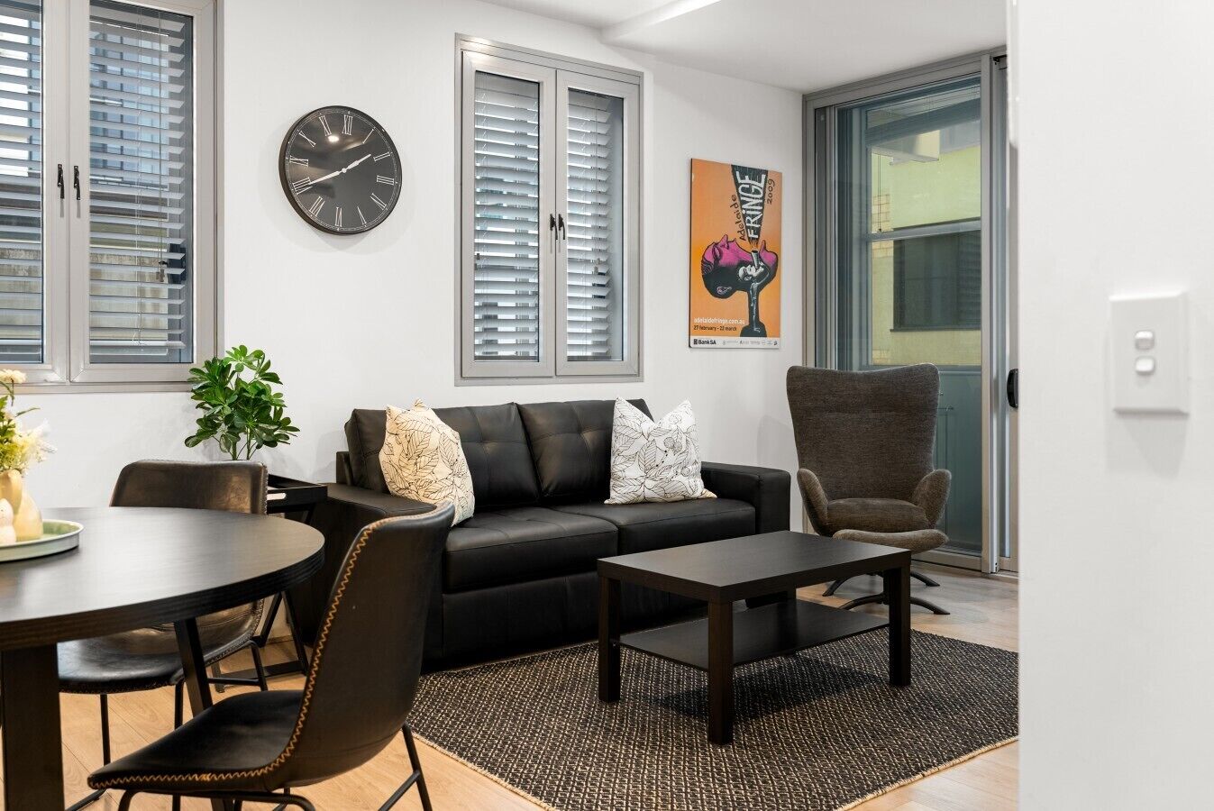 A modern living room with a black leather sofa, a round table, and a brown armchair. There is a clock on the wall and a poster on the wall.
