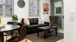 Een moderne woonkamer met een zwarte leren bank, een ronde tafel en een bruine fauteuil. Er hangt een klok aan de muur en een poster.