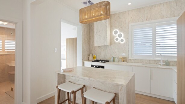 Una cocina moderna con encimera de mármol, dos taburetes de madera, una lámpara colgante y un amplio ventanal con persianas.
