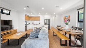 A modern kitchen with white cabinets, a stainless steel refrigerator, and a dining area with a wooden table set for four. - Paradise Palms—Modern Coastal Escape with Backyard (Aldinga Beach)