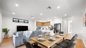 A modern living space with a dining area, grey sofa, wooden dining table, and a kitchen in the background. - Paradise Palms—Modern Coastal Escape with Backyard (Aldinga Beach)