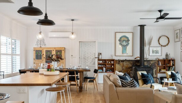 A modern kitchen with a wooden island, bar stools, and a dining table. There is a living area with a sofa and a fireplace.