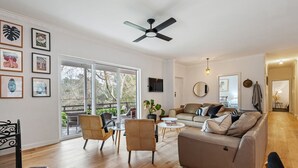 A spacious living room with a sofa, chairs, and a television. There is a ceiling fan and a large window providing natural light.