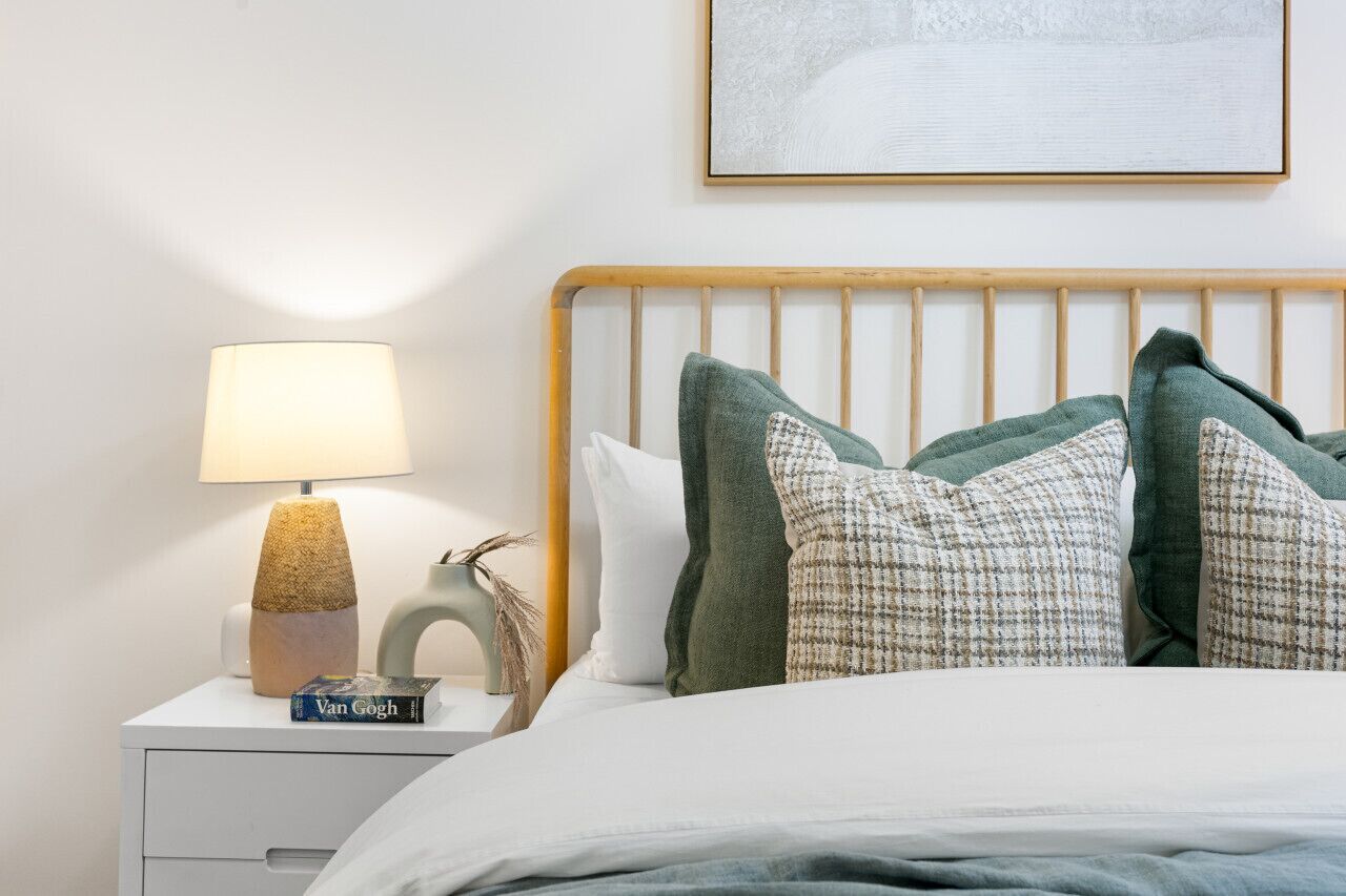 A neatly made bed with a wooden headboard, a bedside lamp, a book titled 'Van Gogh', and a framed picture on the wall.