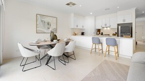 A modern kitchen with white cabinetry, a dining area with white chairs, and a bar with stools. - Mawson Lakes Retreat—A Peaceful and Prime Location (Mawson Lakes)