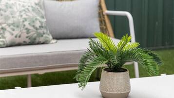 Une table blanche avec une plante en pot au premier plan, un canapé en osier avec des coussins à l’arrière-plan, et un mur vert derrière le canapé.