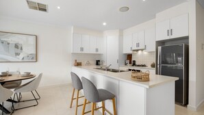 A modern kitchen with white cabinets, a stainless steel refrigerator, and a central island with bar stools. - Mawson Lakes Retreat—A Peaceful and Prime Location (Mawson Lakes)