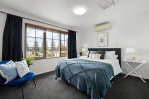 A bedroom with a large window, a bed with a blue blanket, a blue chair, and a bedside table with a lamp.