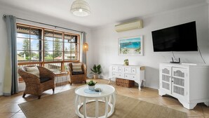 Una sala de estar con un gran ventanal, muebles de mimbre, un aparador blanco y un televisor de pantalla plana.