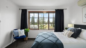 Un dormitorio con un ventanal grande, una cama con colcha azul, un sillón con cojines azules y una mesita de noche con una lámpara.