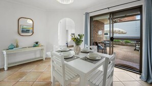 Un comedor con una mesa blanca preparada para cuatro personas, un jarrón con flores y vistas al patio exterior a través de un ventanal.