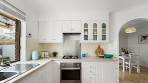 Una cocina con armarios blancos, estufa a gas, lavamanos y un pequeño comedor con mesa y sillas.