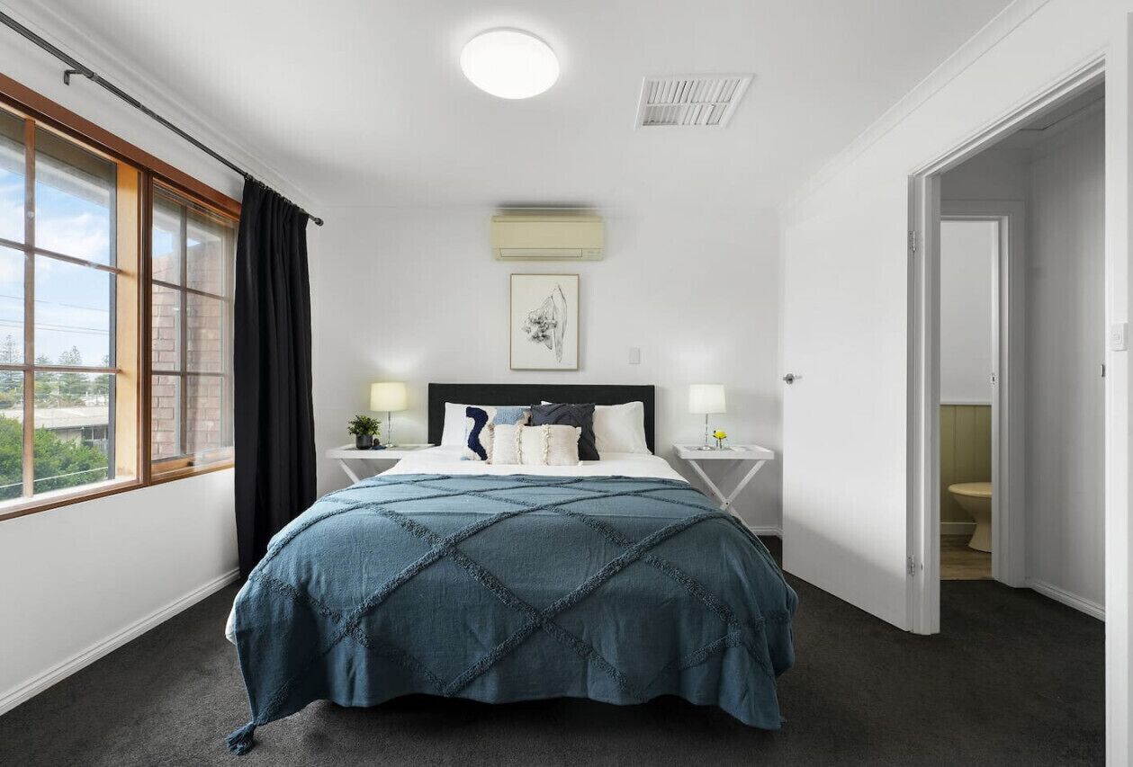 Ein Schlafzimmer mit einem großen Bett, einem Fenster mit Vorhängen, einem Nachttisch mit Lampe und einer Tür zu einem Badezimmer.