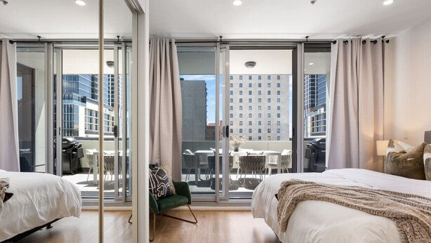 A modern hotel room with two beds, a sliding glass door to a balcony, and a view of the cityscape.