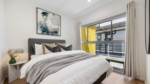 A modern bedroom with a large bed, a nightstand with a lamp, a painting on the wall, and a view of a balcony through a window.