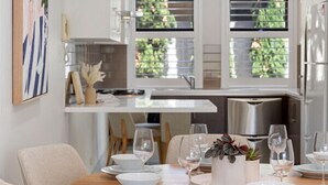 A modern kitchen with white cabinets, a wooden dining table set with white plates and glasses, and a view of the outdoors through large windows.