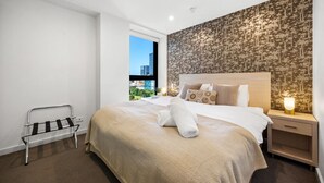 A hotel room with a large bed, a bedside table with a lamp, a window with a view of buildings, and a wall with a patterned wallpaper. - The Balfours Haven - City Views & Balcony (Adelaide)