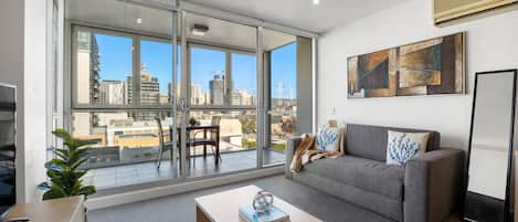 A modern living room with a grey sofa, a wooden coffee table, and a city view through large sliding glass doors.