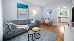 A modern living room with a grey sofa, a coffee table, a dining area with white chairs, a round mirror, and a kitchen visible through an open door. - Brighton Breeze Retreat—Idyllic Beachside Stay (Brighton)