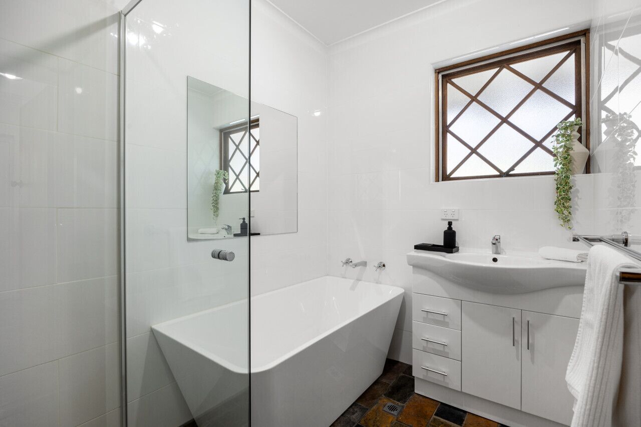 A modern bathroom with a glass shower enclosure, a freestanding bathtub, a sink with a window above it, and white cabinetry.