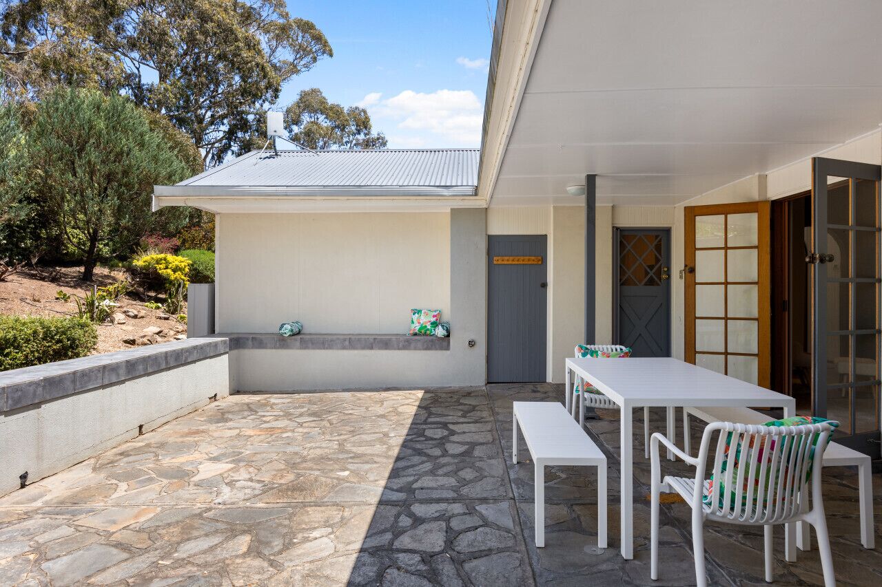 A covered outdoor patio with stone flooring, white furniture, and a view of the surrounding landscape.