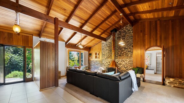 A spacious living area with a large black sectional sofa, wooden beams, stone walls, and a view of greenery outside. - Amber Wisper—In the Hills with Pool & Tennis Court (Paechtown)