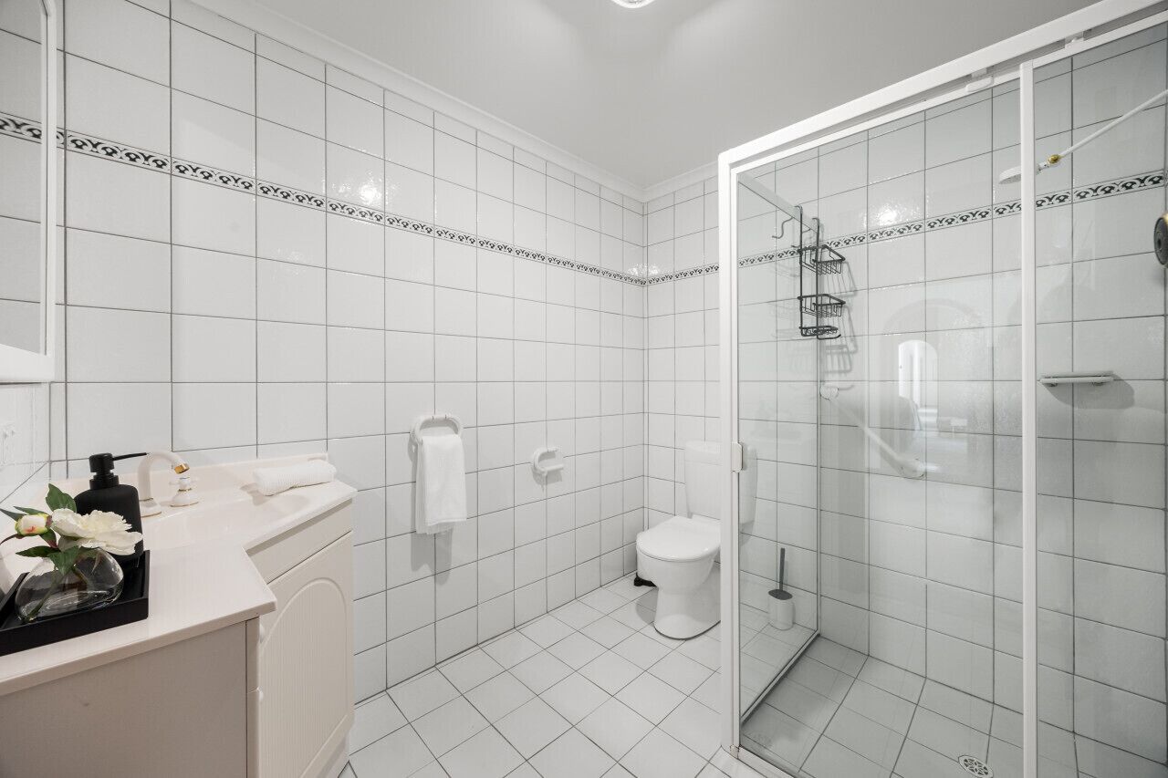 Un baño moderno con azulejos blancos, cabina de ducha de vidrio, inodoro blanco y tocador con espejo y artículos de aseo.