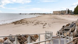 Une plage avec une jetée qui s’avance dans l’eau, une côte rocheuse et, au loin, un bâtiment doté de nombreux balcons.