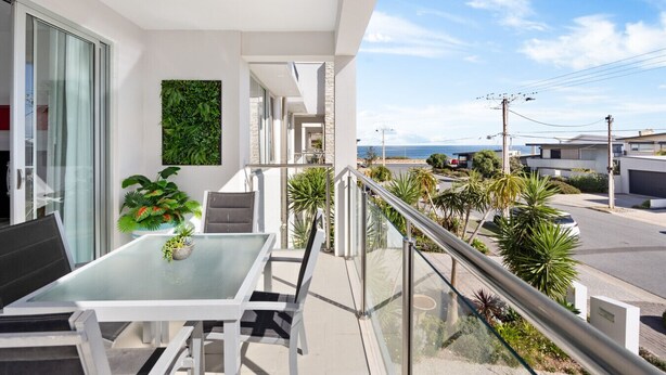 Un balcon avec une table et des chaises en verre, une plante en pot et une vue sur la mer et les bâtiments voisins.