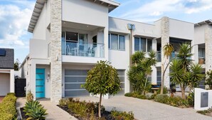 Un immeuble résidentiel moderne à deux étages, doté d’un balcon, d’un garage et d’un jardin bien entretenu où poussent différentes espèces de plantes.