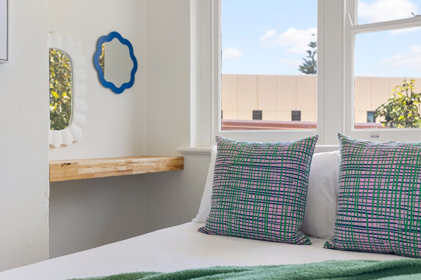 Een kamer met een raamzitje, twee gekleurde kussens, een blauwe decoratieve spiegel en een houten plank.