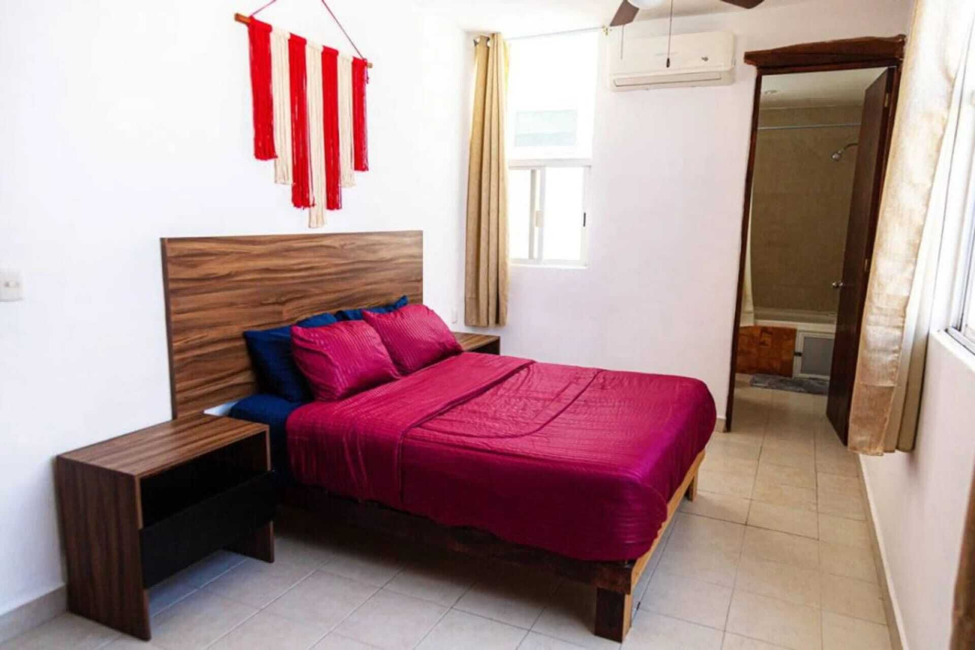 A bedroom with a wooden bed, a nightstand, a red and white wall hanging, and a window with curtains.